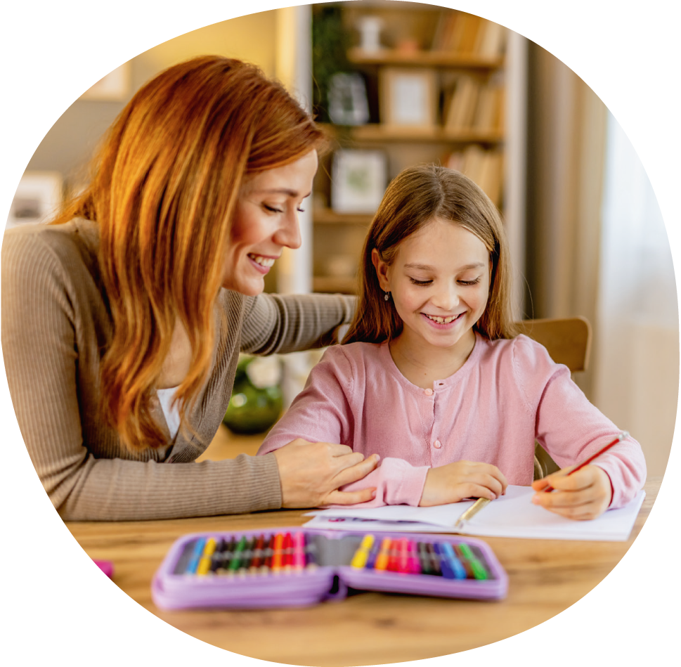 Eine Mutter sitzt mit ihrer Tochter am Esstisch. Sie legt ihren Arm um ihre Tochter. Die Tochter schreibt in ein Heft. Vor ihnen liegt eine Federmappe auf dem Tisch.