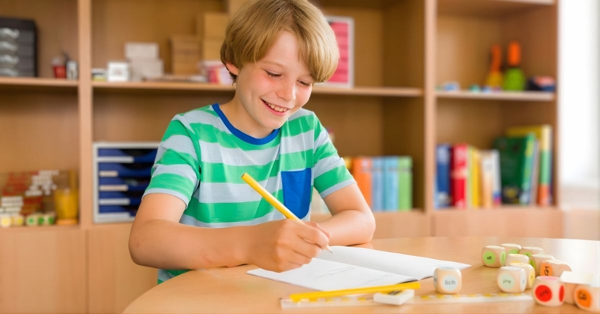 Ein Junge sitzt in der Lerntherapie an einem Schreibtisch und schreibt etwas auf ein Blatt Papier. Daneben liegen Wortbausteinwürfel. Im Hintergrund ist ein Regal zu sehen mit weiteren Hilfsmitteln aus der Lernthterapie.