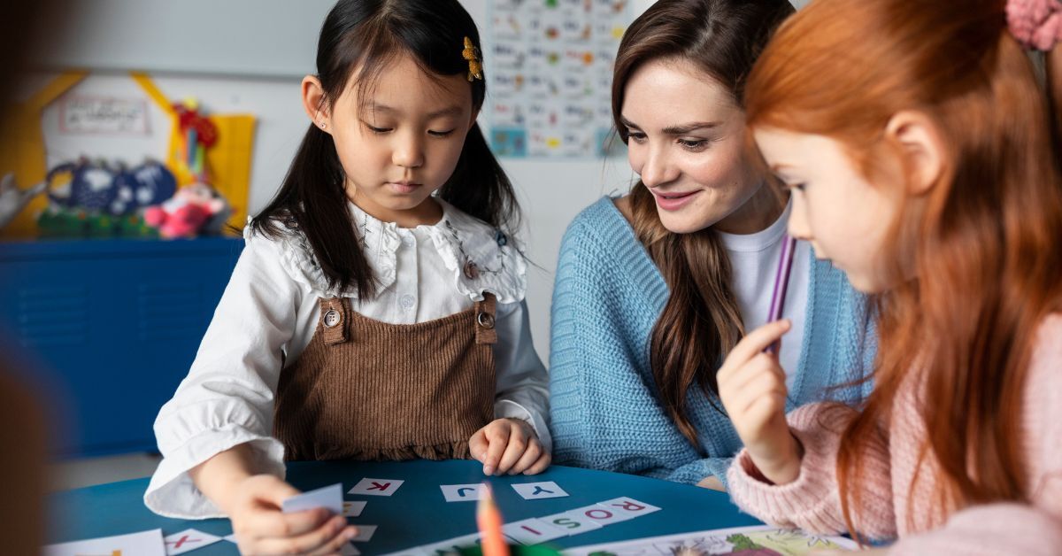 Zwei Schulmädchen sitzen mit ihrer Lehrerin an einem Tisch und legen Buchstabenkarten.